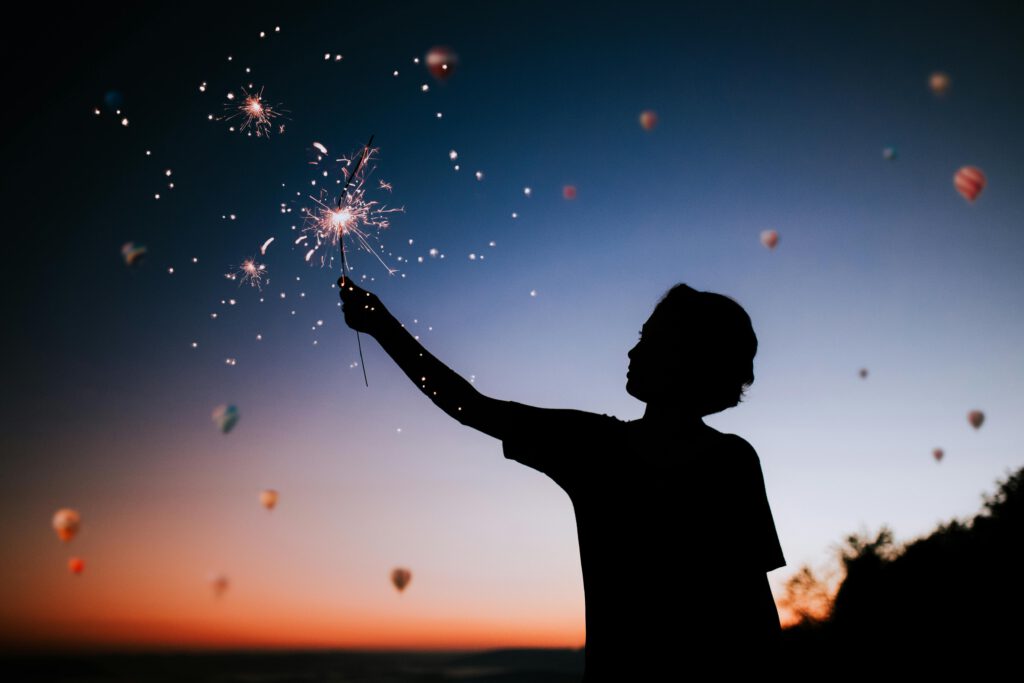Silhouette of a person holding sparklers against a colorful twilight sky.